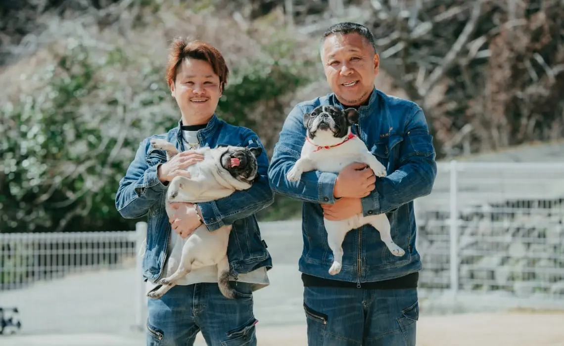 デニム姿の男性2人が犬を抱いて笑顔で並んでいる。背景は屋外のドッグランのような場所。